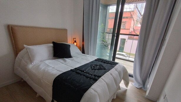 A neatly made bed with a white duvet and black throw, a wooden headboard, and a view of a red building through a large window. - Estudio en Palermo 4 (Buenos Aires)