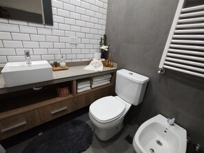 A modern bathroom with a white toilet, a sink, and a bidet. There is a wooden vanity with a mirror above the sink, and a black rug on the floor.