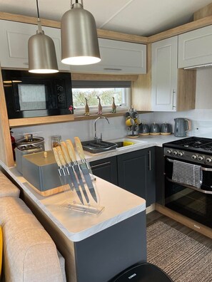 A compact kitchen with a microwave, stove, sink, and a knife block. - KMH Caravan Holidays (Malton)