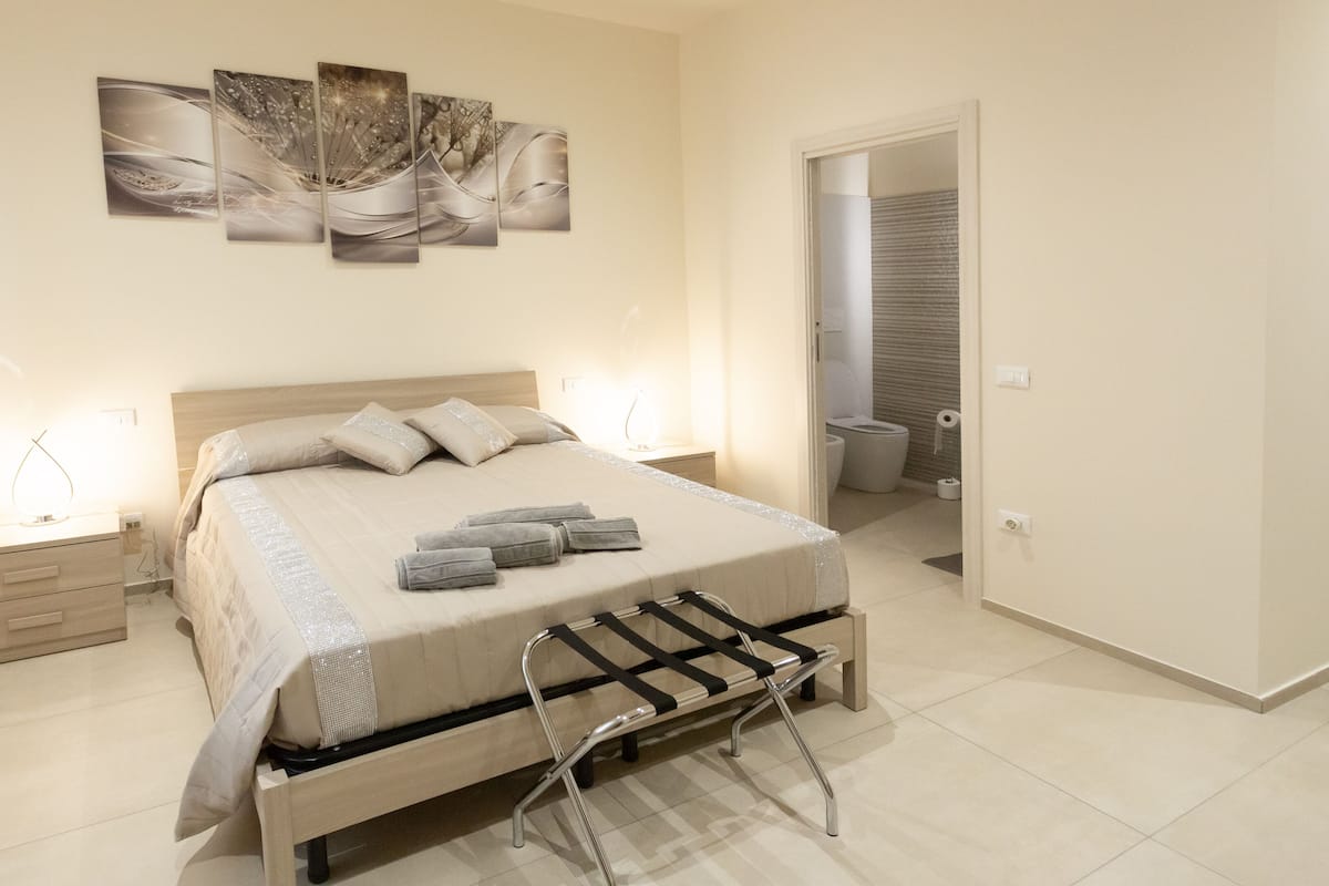 A modern bedroom with a bed, bedside tables, a bench, and a bathroom visible through an open door.
