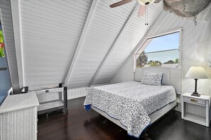 A bedroom with a sloped ceiling, a bed with a patterned bedspread, a nightstand with a lamp, and a window with a view of the outside. - Happy Daze Home, Fire Pit, Gazebo & Vilano Pier (St. Augustine)