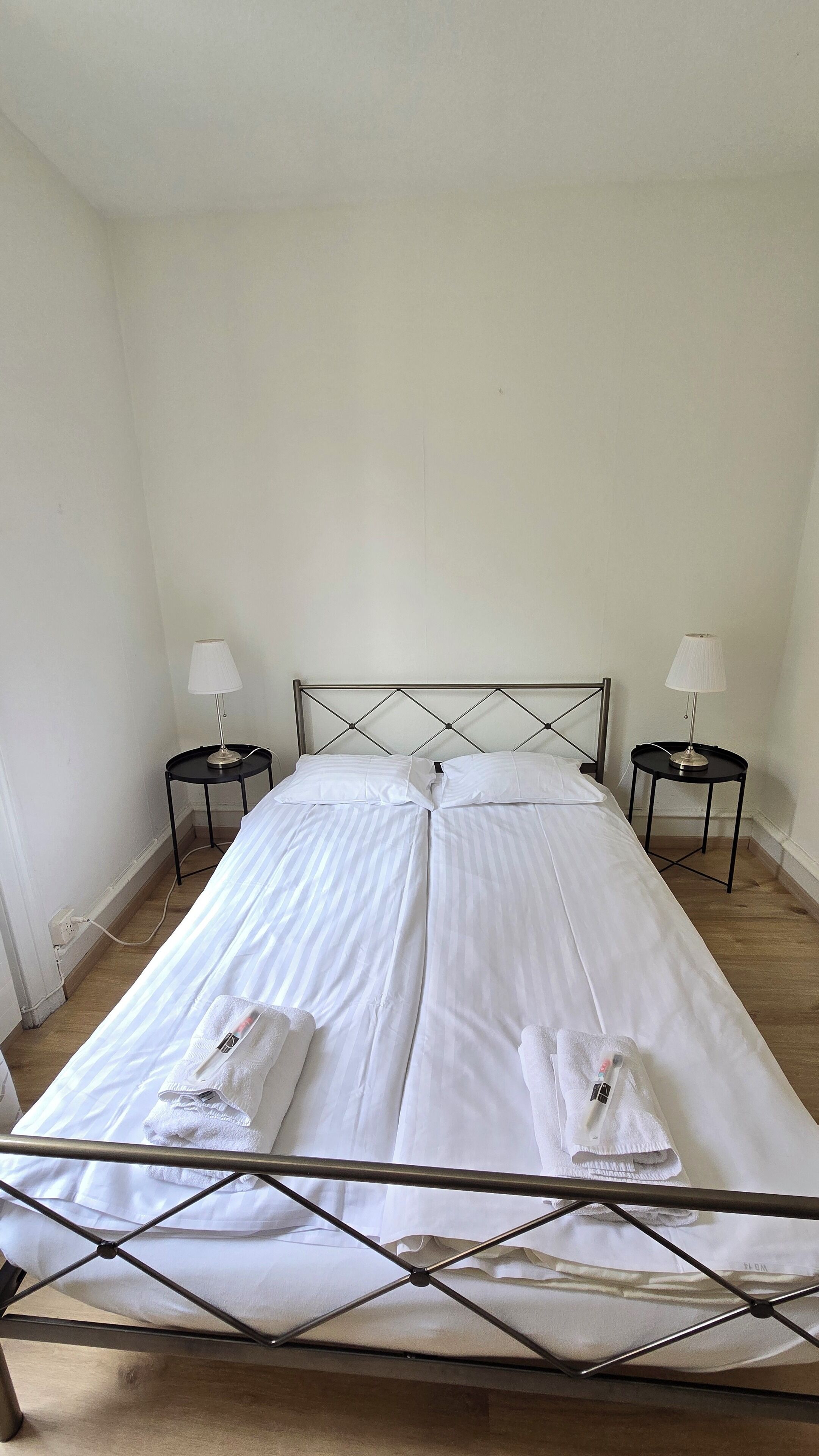 Un lit simple avec un cadre en métal, deux tables de chevet avec des lampes et un mur blanc uni.