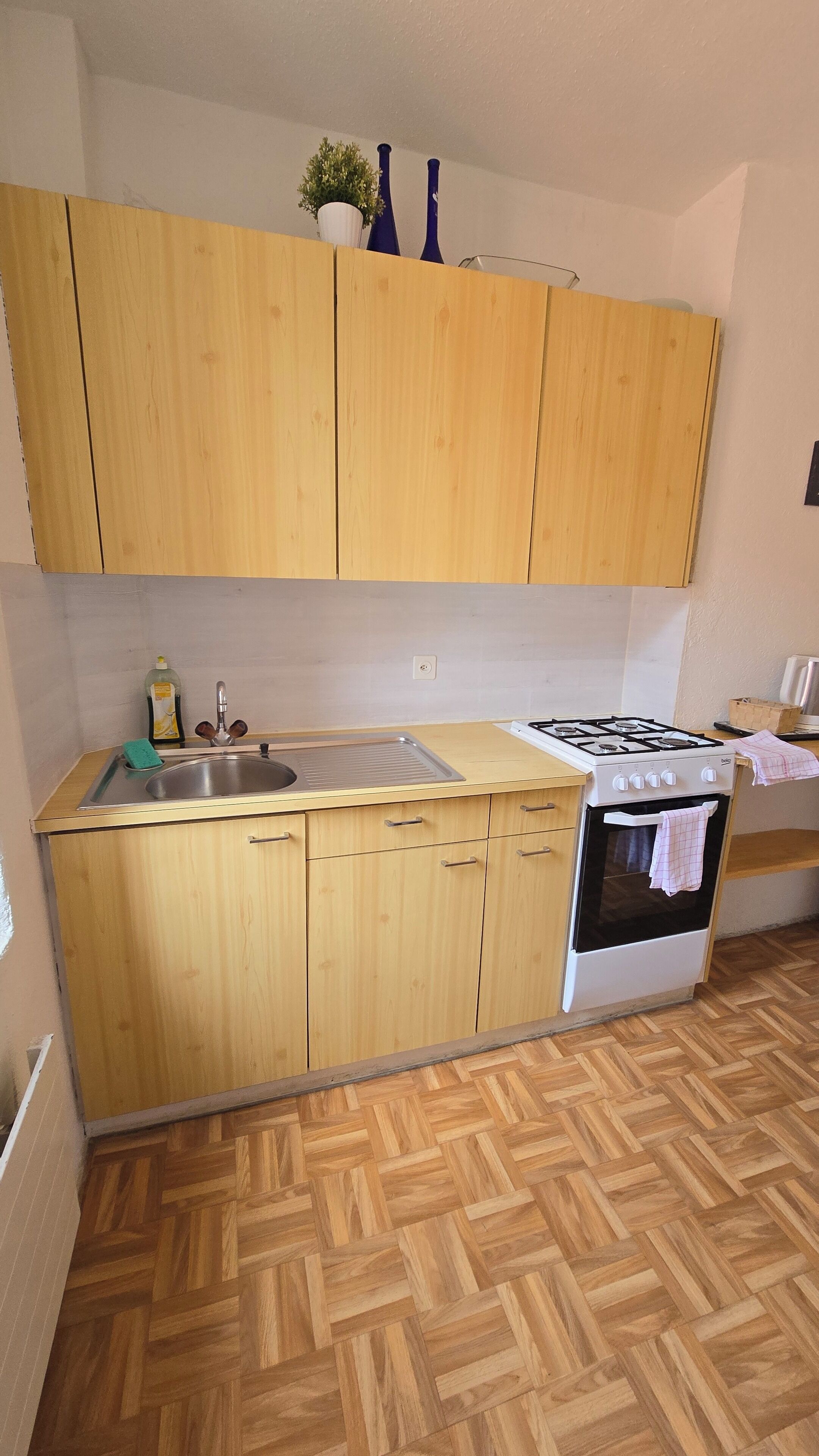 A compact kitchen with wooden cabinets, a stainless steel sink, a gas stove, and a herringbone-patterned wooden floor.