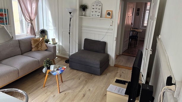 A living room with a sofa, ottoman, and a table. There is a television on a stand and a plant on the wall. - Mi Casa es su Casa (Zürich)