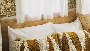 Ein gemütliches Interieur mit einer Wand mit Muster, einem Bett mit weißen und senfgelben Kissen und einem Fenster mit durchsichtigen Vorhängen.