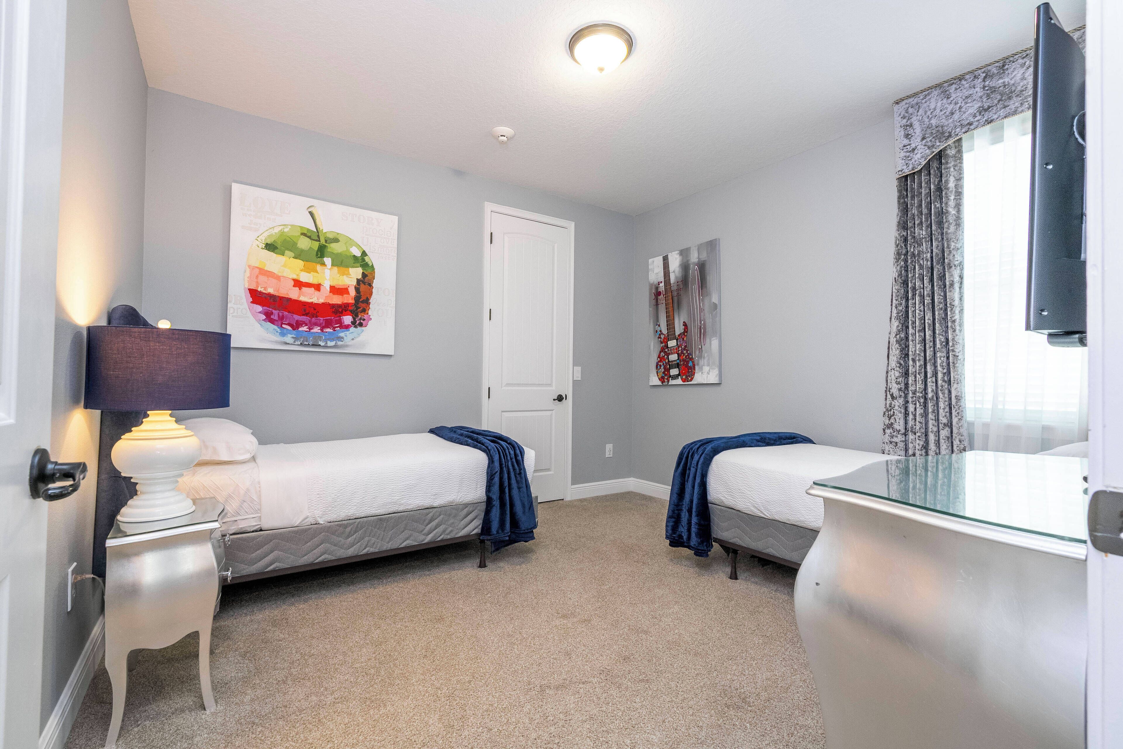 A bedroom with two single beds, a nightstand with a lamp, a wall-mounted TV, and artwork.