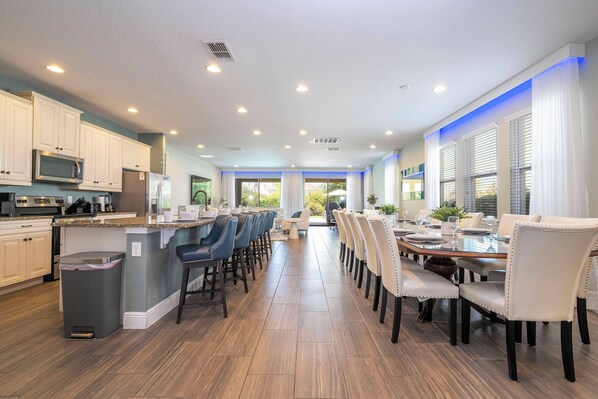 A spacious kitchen with modern appliances, a dining area with a long table and chairs, and a view of the outdoors. - Luxury 9BR Villa - Private Pool, Spa, Game Room - Free Pool Heat in Aug - Oct! (Kissimmee)