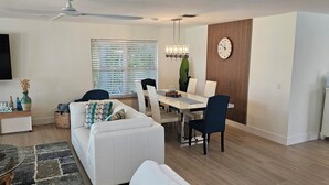 Un soggiorno moderno con televisore a schermo piatto, un'area pranzo con tavolo e sedie, un divano con cuscini decorati e un pannello decorativo a parete.