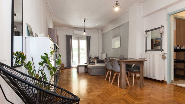 A spacious living area with a wooden floor, a dining table with chairs, a black chair, and a potted plant. - Modern 2 Bd Kipseli Hideaway Patio Apt (Athens)
