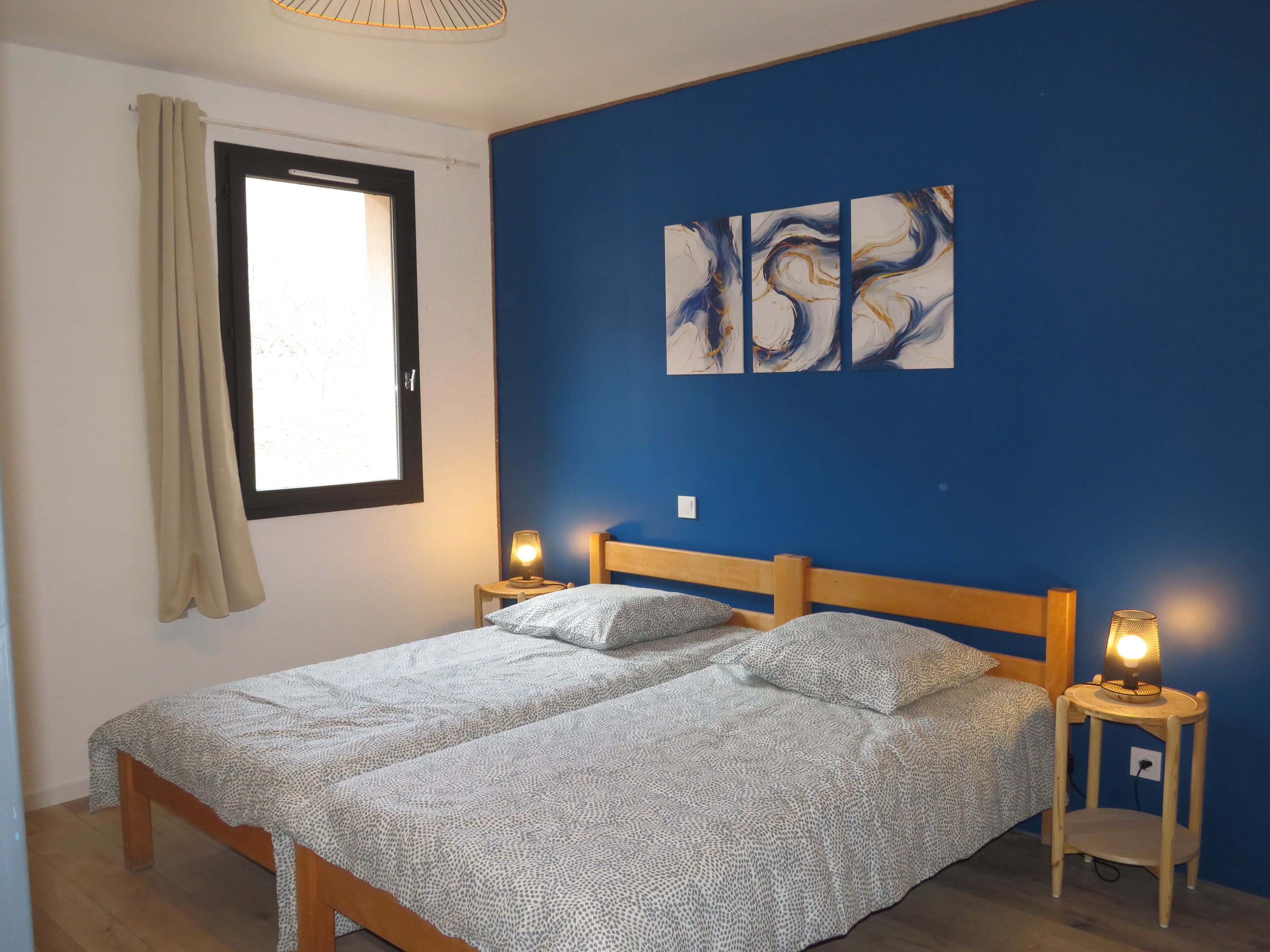 Une chambre à coucher avec un lit double, deux lampes de chevet, une fenêtre avec des rideaux et des œuvres d’art abstraites accrochées au mur.