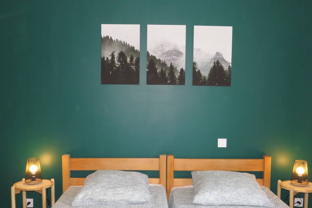 A room with two beds, a green wall, and three framed pictures of mountains.