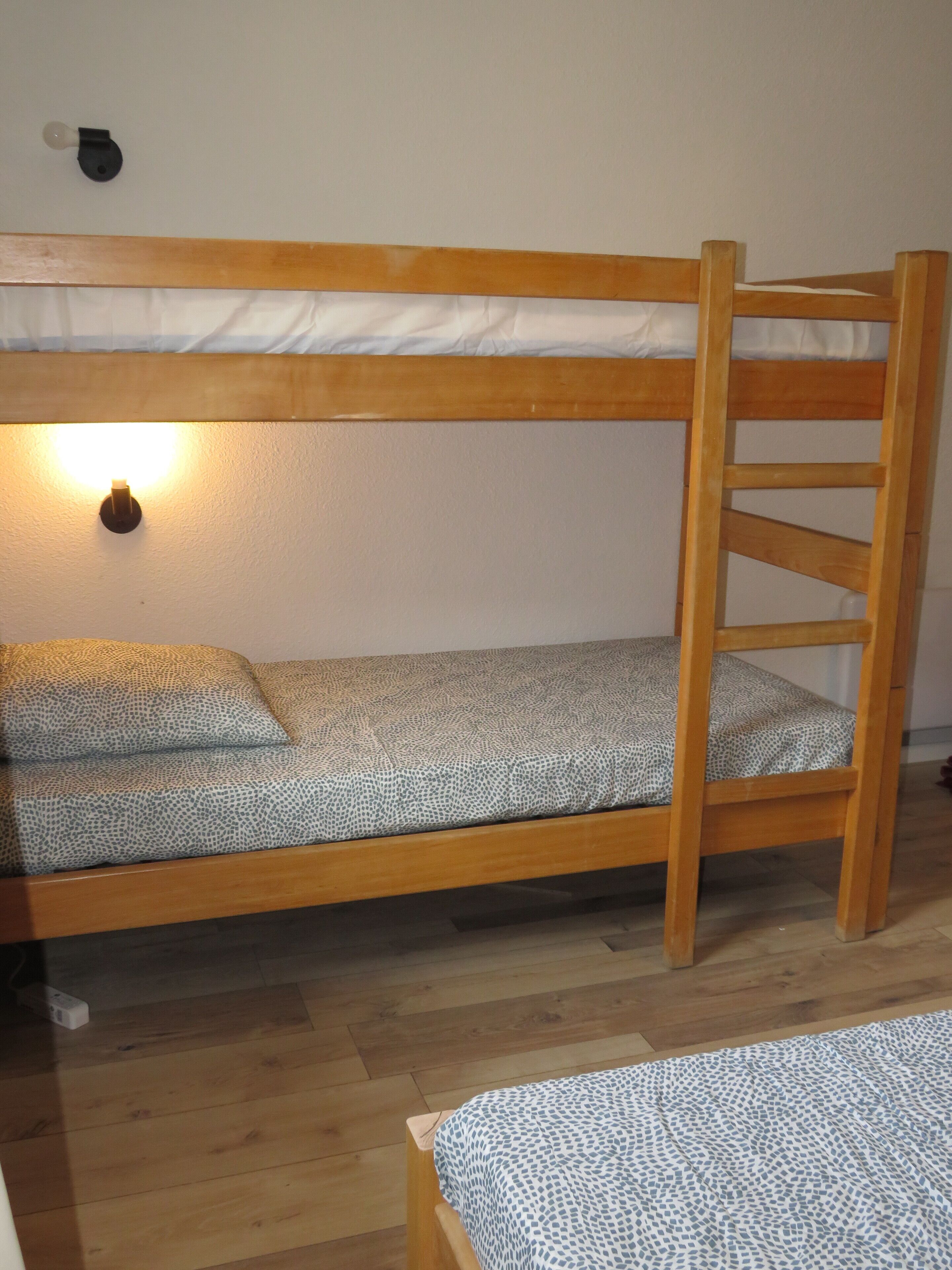 Una cama individual superpuesta con escalera, una lámpara fijada a la pared y una colcha estampada.