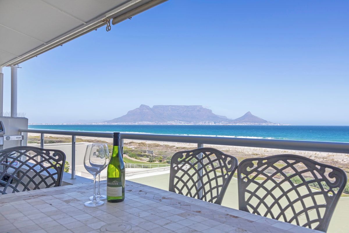 A table with a bottle of wine and a glass, overlooking a beach and a mountain range.