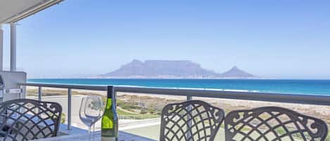 A table with a bottle of wine and a glass, overlooking a beach and a mountain range.