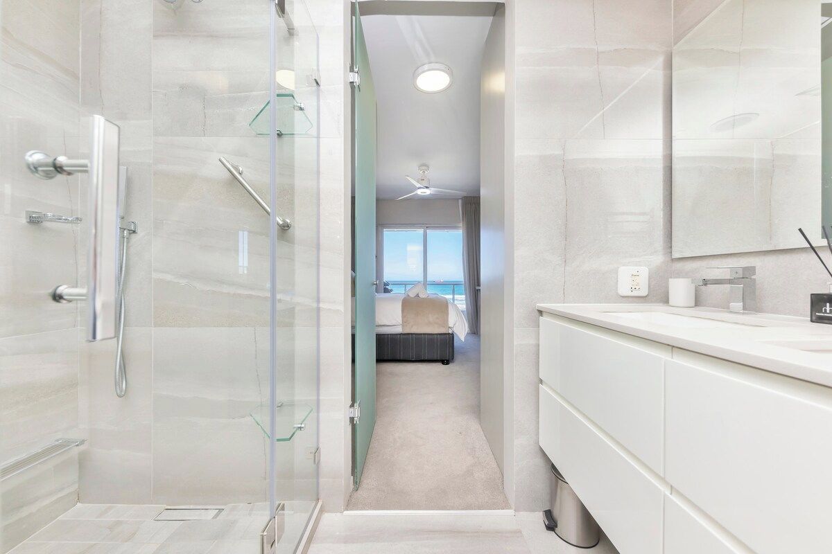 Een moderne badkamer met een douche met glazen wand, een grote spiegel en uitzicht op de oceaan door een raam.