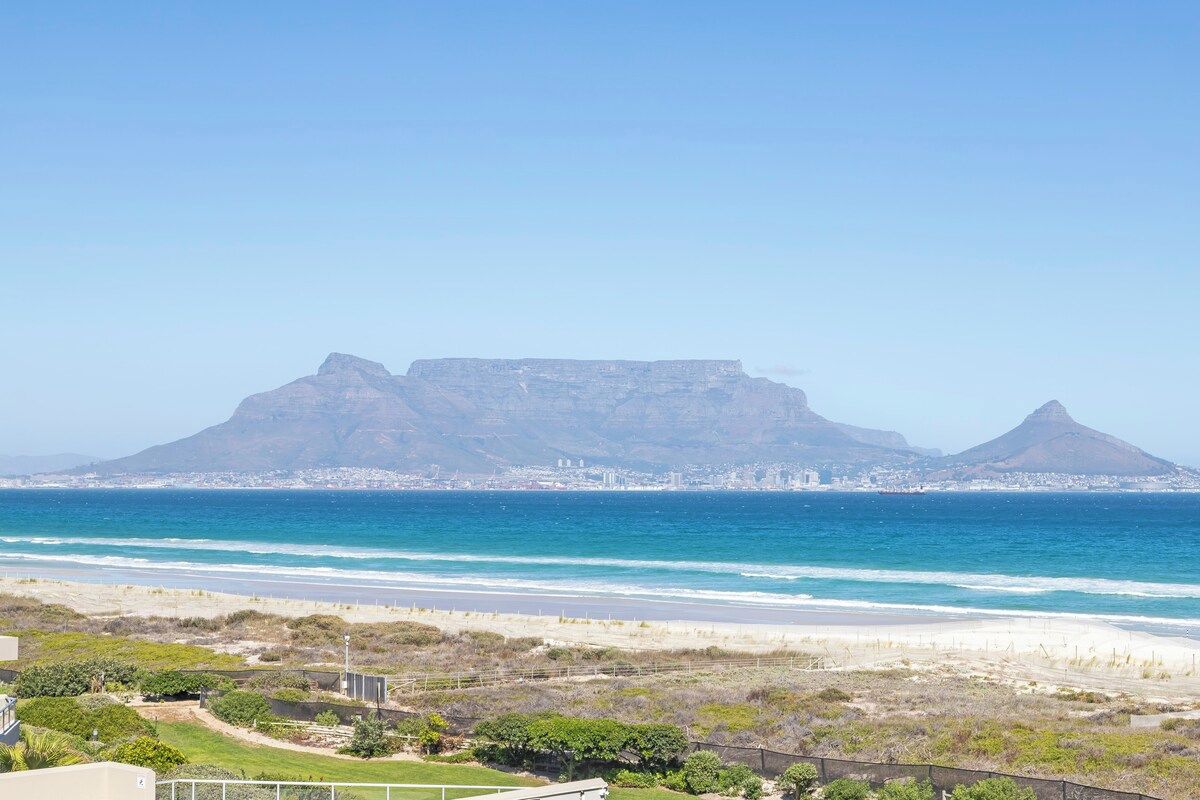 Un paisaje costero con una montaña destacada al fondo, una playa en primer plano y un paisaje urbano al otro lado del agua.