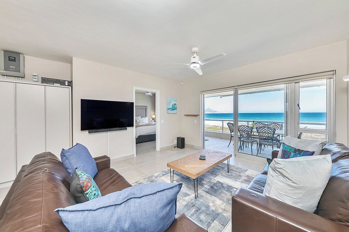 A living room with a large TV, a ceiling fan, and a view of the ocean through large windows.