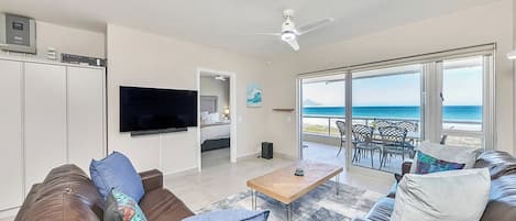 A living room with a large TV, a ceiling fan, and a view of the ocean through large windows.