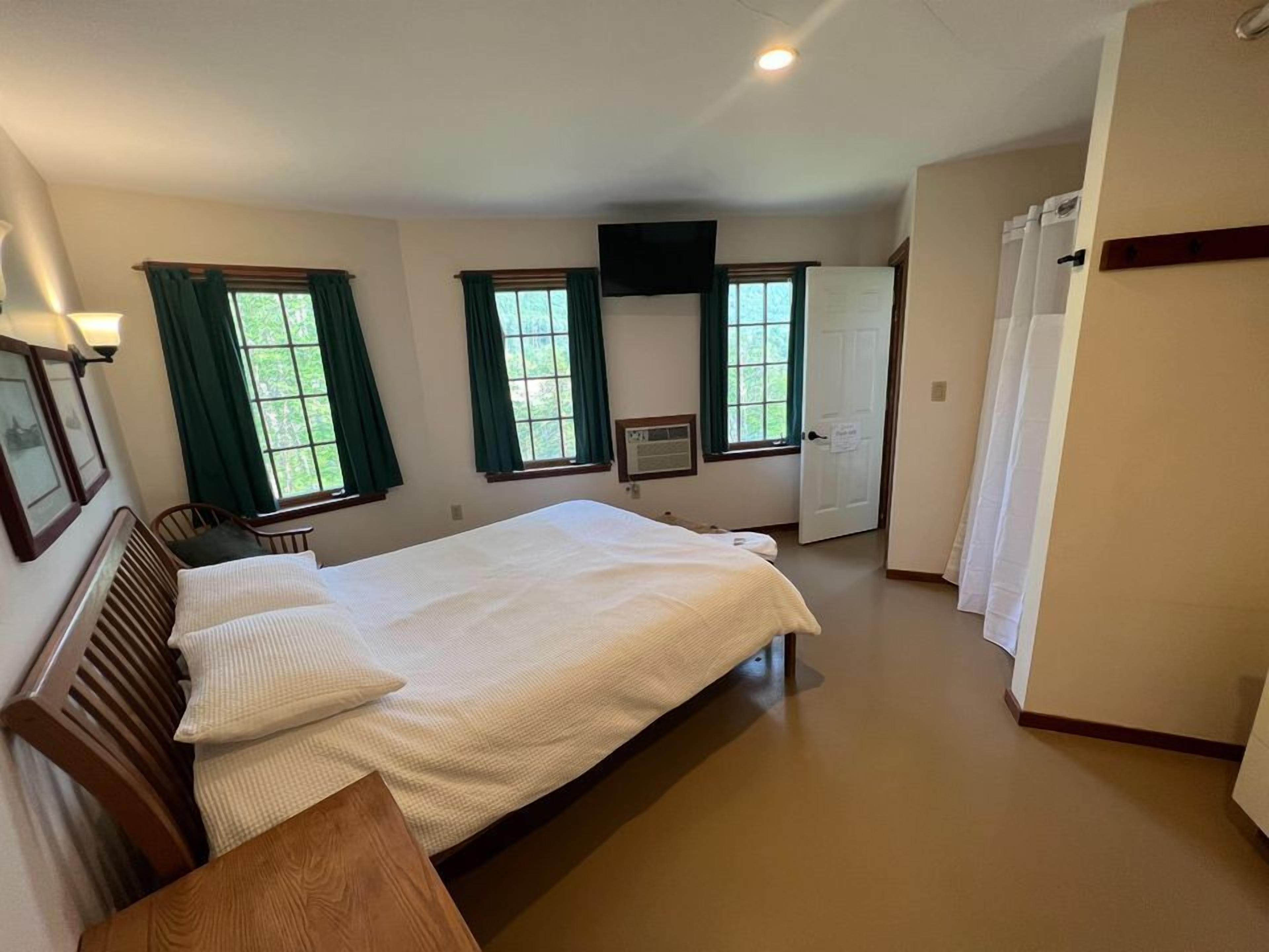 A bedroom with a large bed, two windows with curtains, a flat-screen TV mounted on the wall, and a door with a sign.