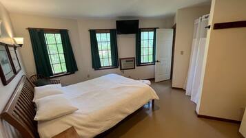A bedroom with a large bed, two windows with curtains, a flat-screen TV mounted on the wall, and a door with a sign.