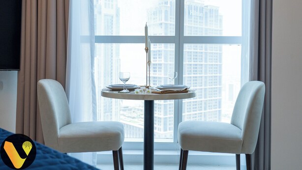 A hotel room with a round dining table set for two, two white chairs, a TV mounted on the wall, and a view of high-rise buildings through large windows.