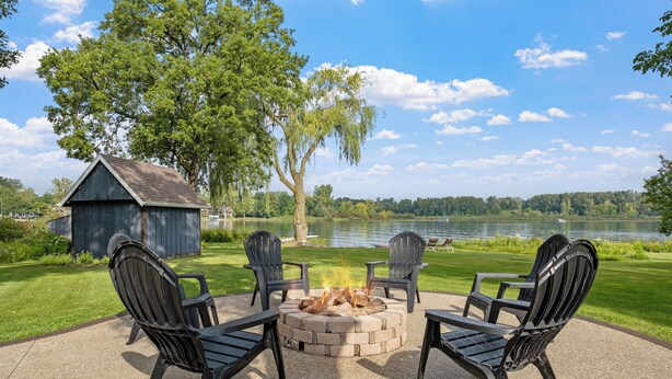 A fire pit with four Adirondack chairs around it, set on a concrete patio with a view of a lake and trees. - Murray Lake Waterfront Retreat w/ Dock, Patio & Fire Pit (Lowell)