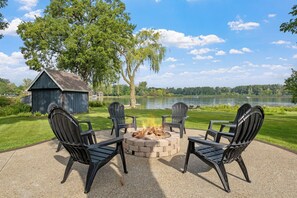 Un foyer extérieur entouré de quatre chaises Adirondack, installé sur une terrasse en béton offrant une vue sur un lac et des arbres.