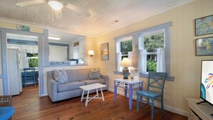 Una sala de estar acogedora con un sofá gris, una mesa de centro de madera y un sillón azul. Se ve un comedor con una mesa y sillas al fondo.
