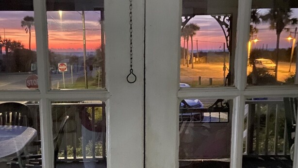 Vista a través de una ventana con una cadena colgando en el centro, que da a una calle con palmeras y un atardecer.