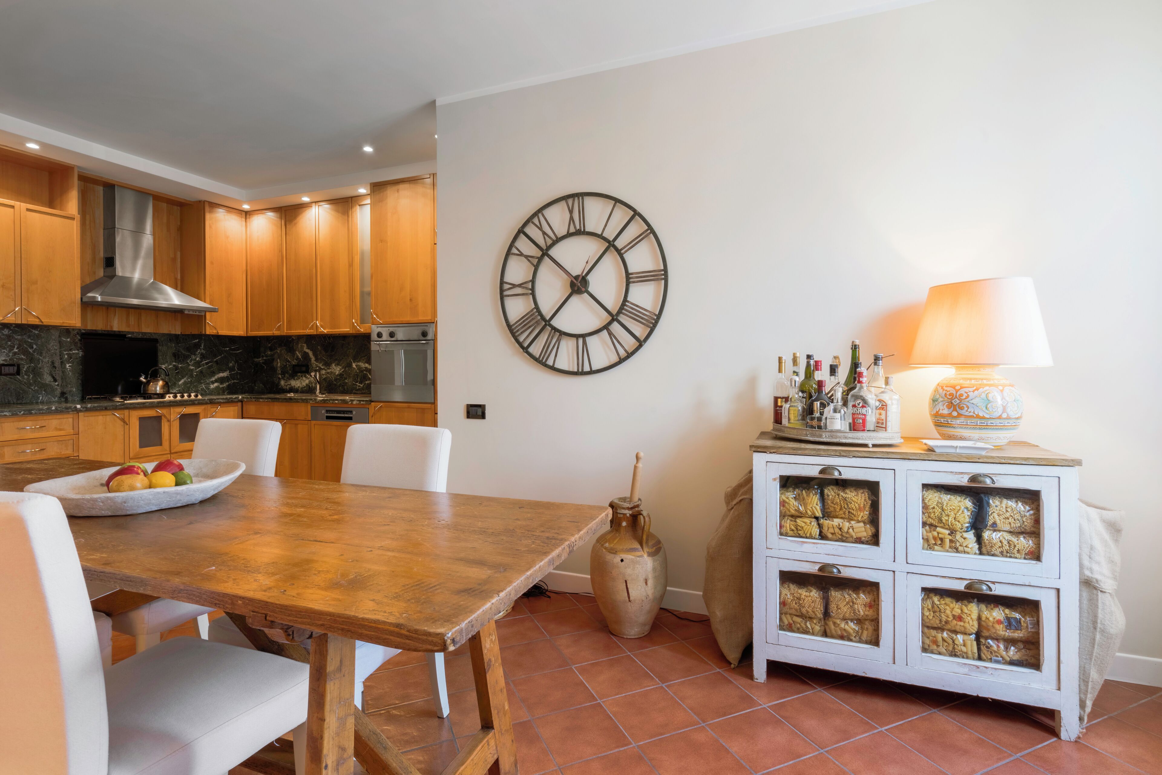 A kitchen with wooden cabinets, a marble countertop, a large wall clock, a dining table with chairs, and a cabinet with glass doors.