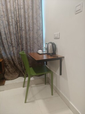 A small room with a wooden table, a green chair, a kettle, and a curtain with a tree pattern. - Treebo Park Palace (Guwahati)