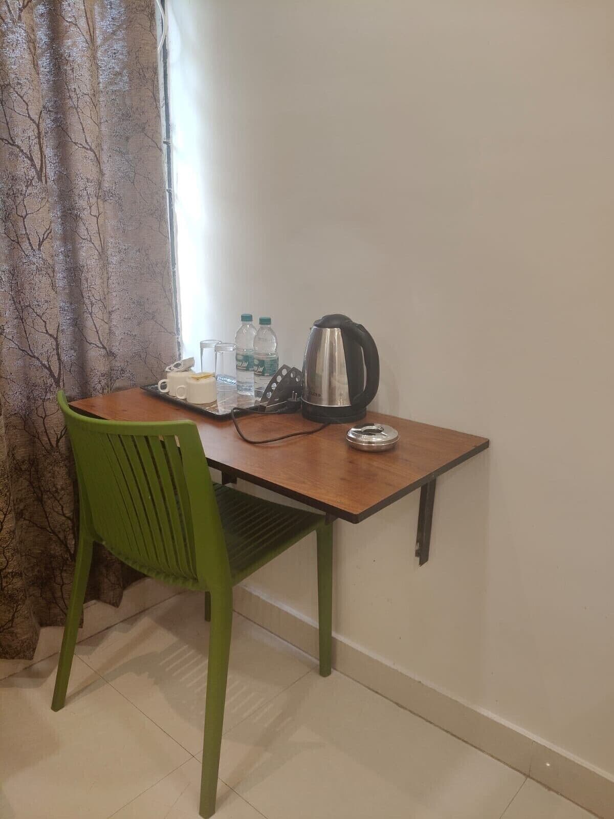 A small, simple room with a wooden desk, a green chair, a kettle, and two bottles of water.