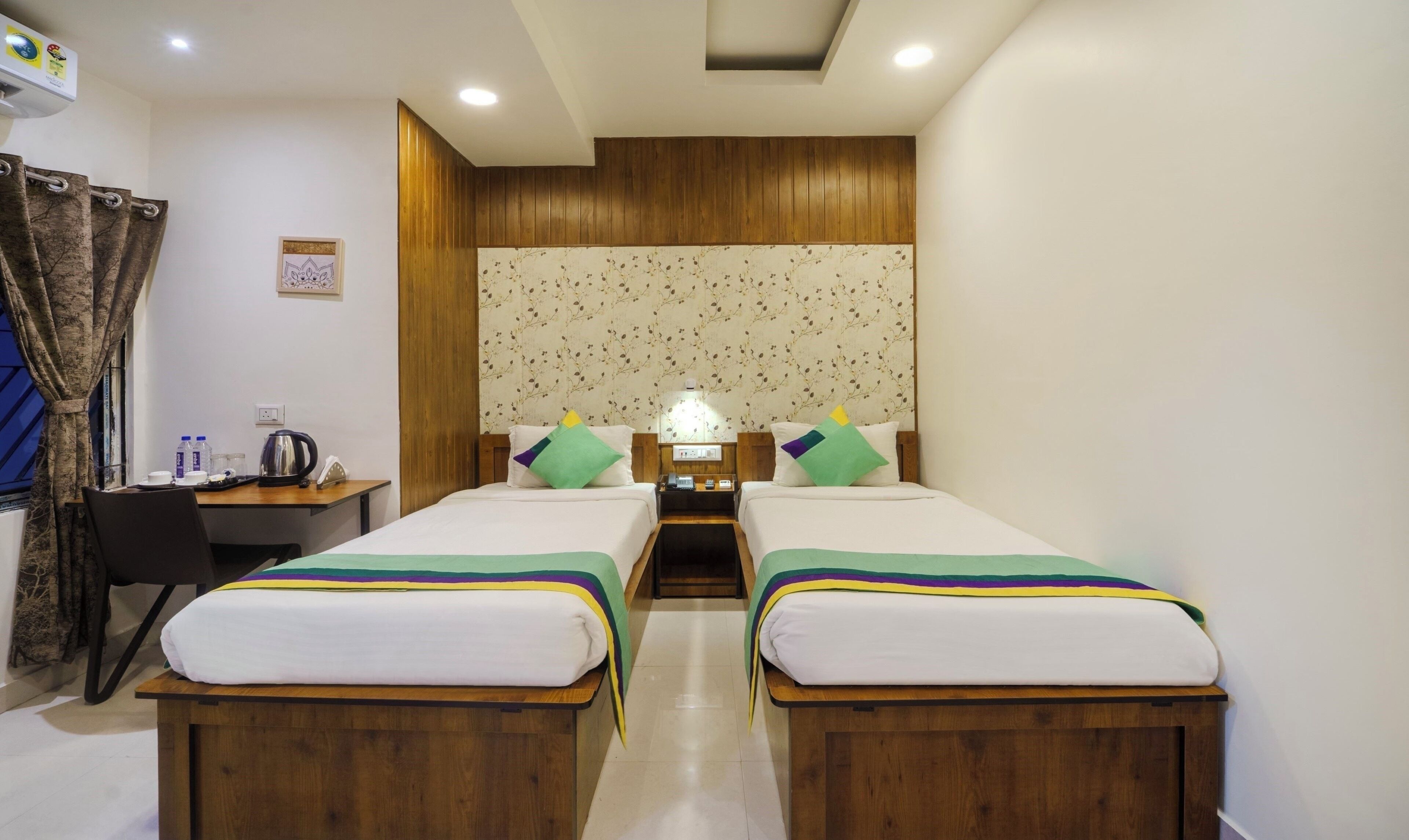 Habitación de hotel con dos camas individuales, un escritorio con una silla, una mesita con un hervidor de agua y un aire acondicionado instalado en la pared.