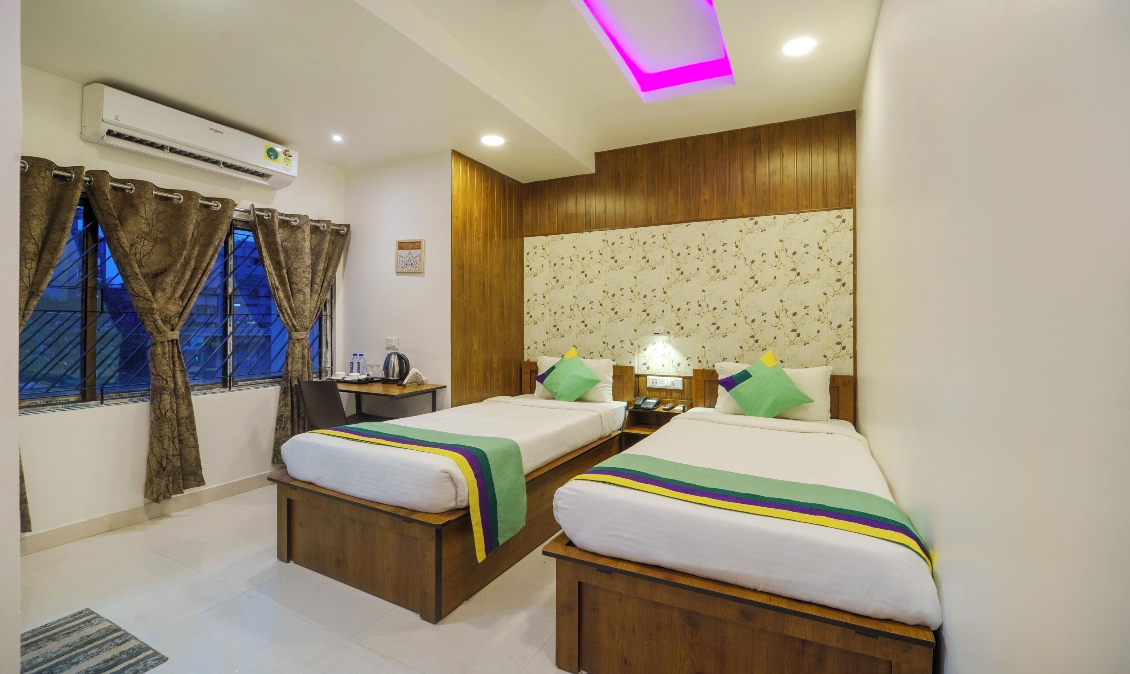 Habitación de hotel con dos camas, cabecero de madera, unidad de aire acondicionado, mesitas de noche y una ventana con cortinas.