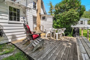Une terrasse en bois avec une chaise et une table, un petit porche avec une fenêtre, et un arbre en arrière-plan.