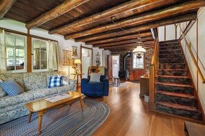Uma sala de estar aconchegante com teto de madeira, um sofá, uma poltrona azul, uma mesa de centro de madeira e uma escada com corrimão de madeira.