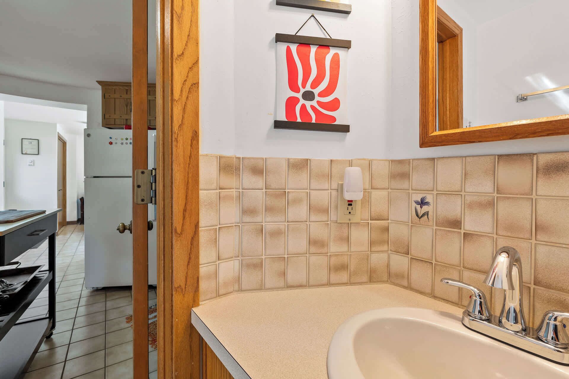 Une salle de bain avec un mur recouvert de carreaux beiges, un lavabo avec un robinet chromé, un distributeur de savon liquide et un miroir encadré en bois.