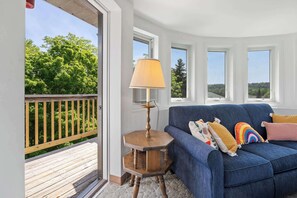 A room with a blue sofa, a wooden side table with a lamp, and a view of greenery through large windows.