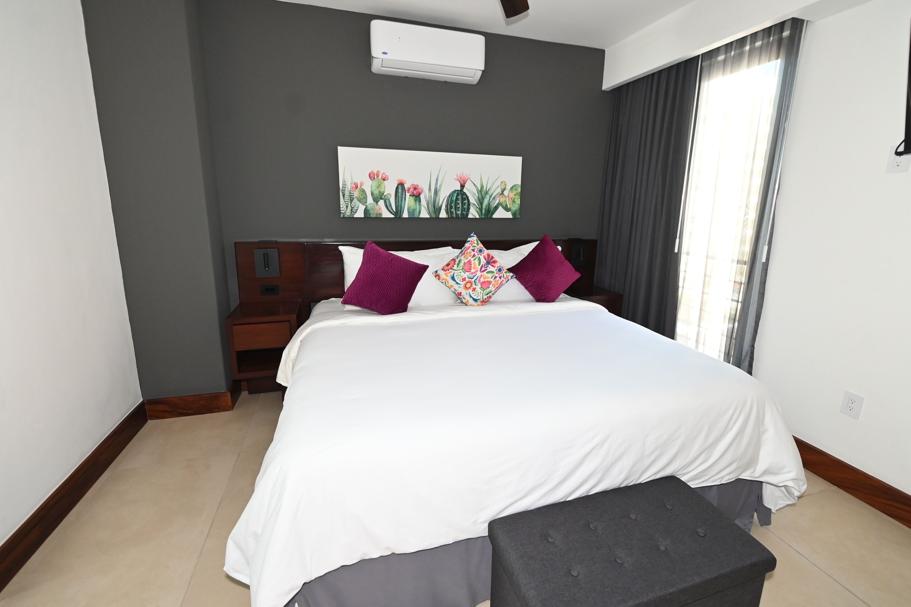 Ein Hotelzimmer mit einem großen Bett, einem Nachttisch, einer grauen gepolsterten Bank, einer Wand-Klimaanlage und einem mit Vorhängen versehenen Fenster.