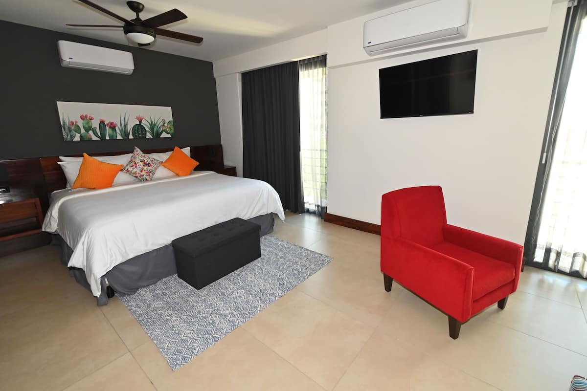 A bedroom with a bed, a red armchair, a ceiling fan, an air conditioning unit, and a window with curtains.