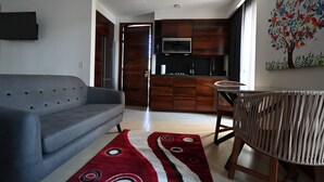 Una sala moderna con un sofá gris, una mesa de comedor con una silla de fibras naturales, una zona de cocina con armarios de madera y un televisor montado en la pared.