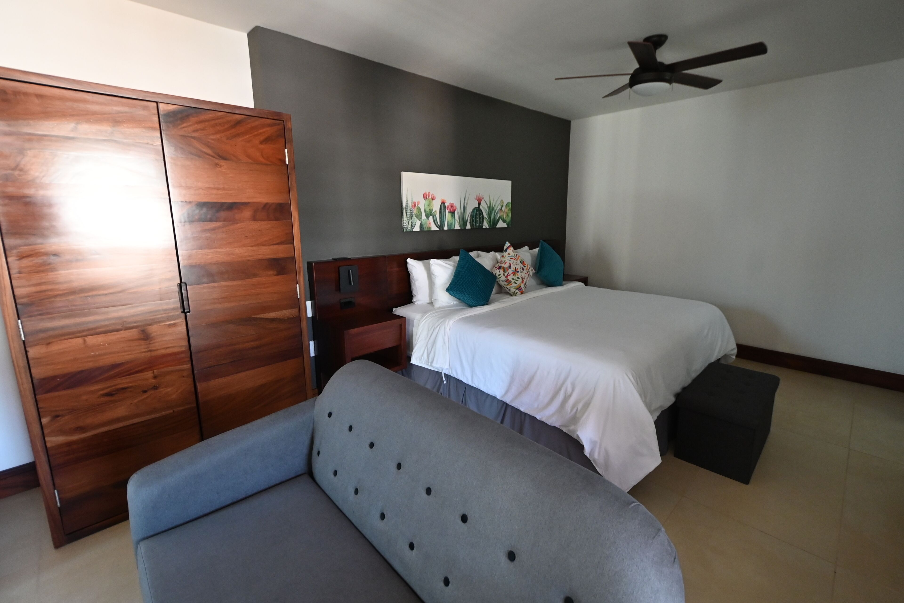 Quarto com cama, sofá cinza, guarda-roupa de madeira, ventoinha no teto e uma obra de arte pendurada na parede.
