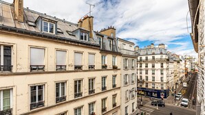 A multi-story building with a row of windows on the ground floor, a visible storefront, and a street in the foreground. - GuestReady - Parisian haven near attractions (Paris)