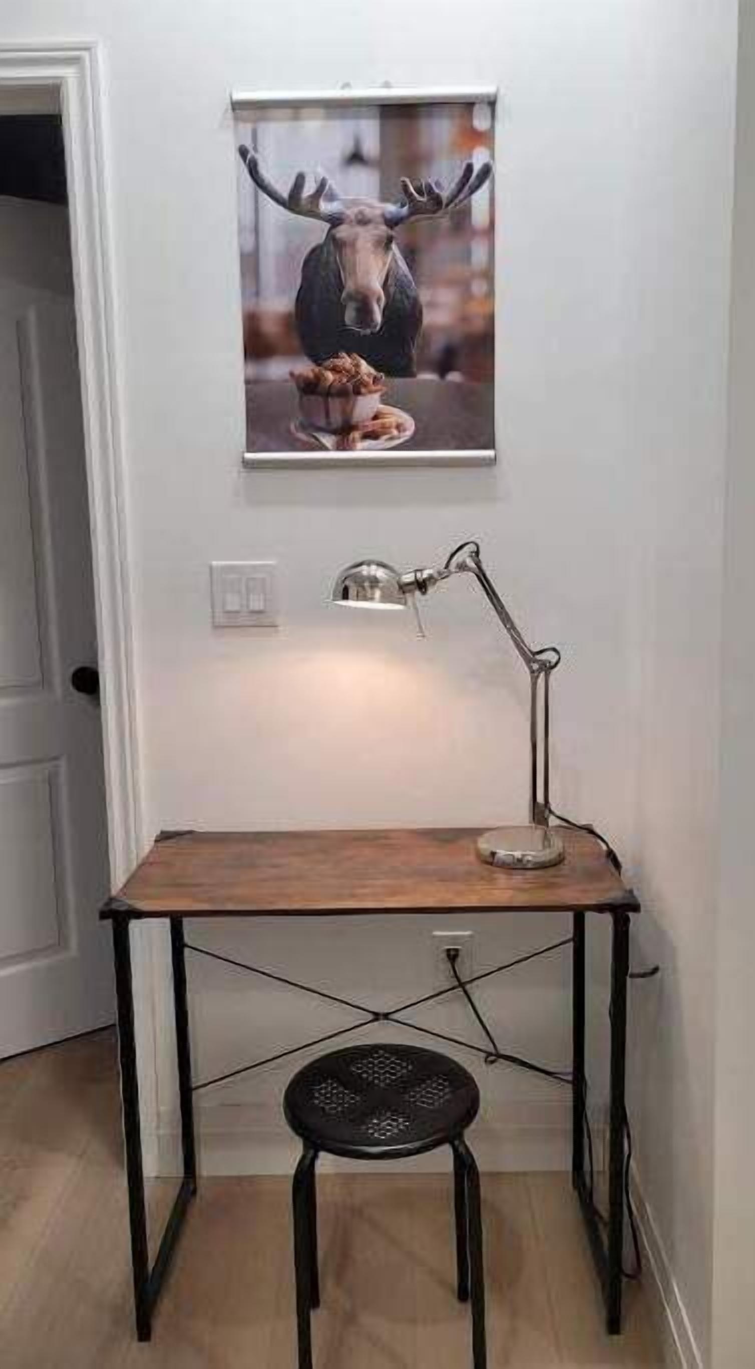 Un petit bureau en bois, agrémenté d’une lampe en métal et d’une chaise noire, est placé dans une pièce aux murs blancs.