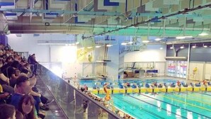 A swimming pool with multiple lanes, spectators seated on bleachers, and a few people standing by the pool.