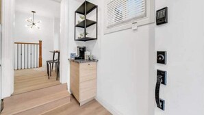 A hallway with a wooden floor, a desk with a coffee maker, a chair, and a wall-mounted shelf.