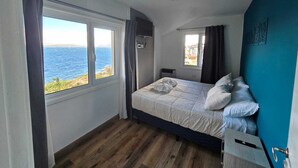 A bedroom with a large window offering a view of the sea, a bed with white bedding, a blue accent wall, and a small desk with a chair. - Los Dones Departamentos DAT (San Carlos de Bariloche)