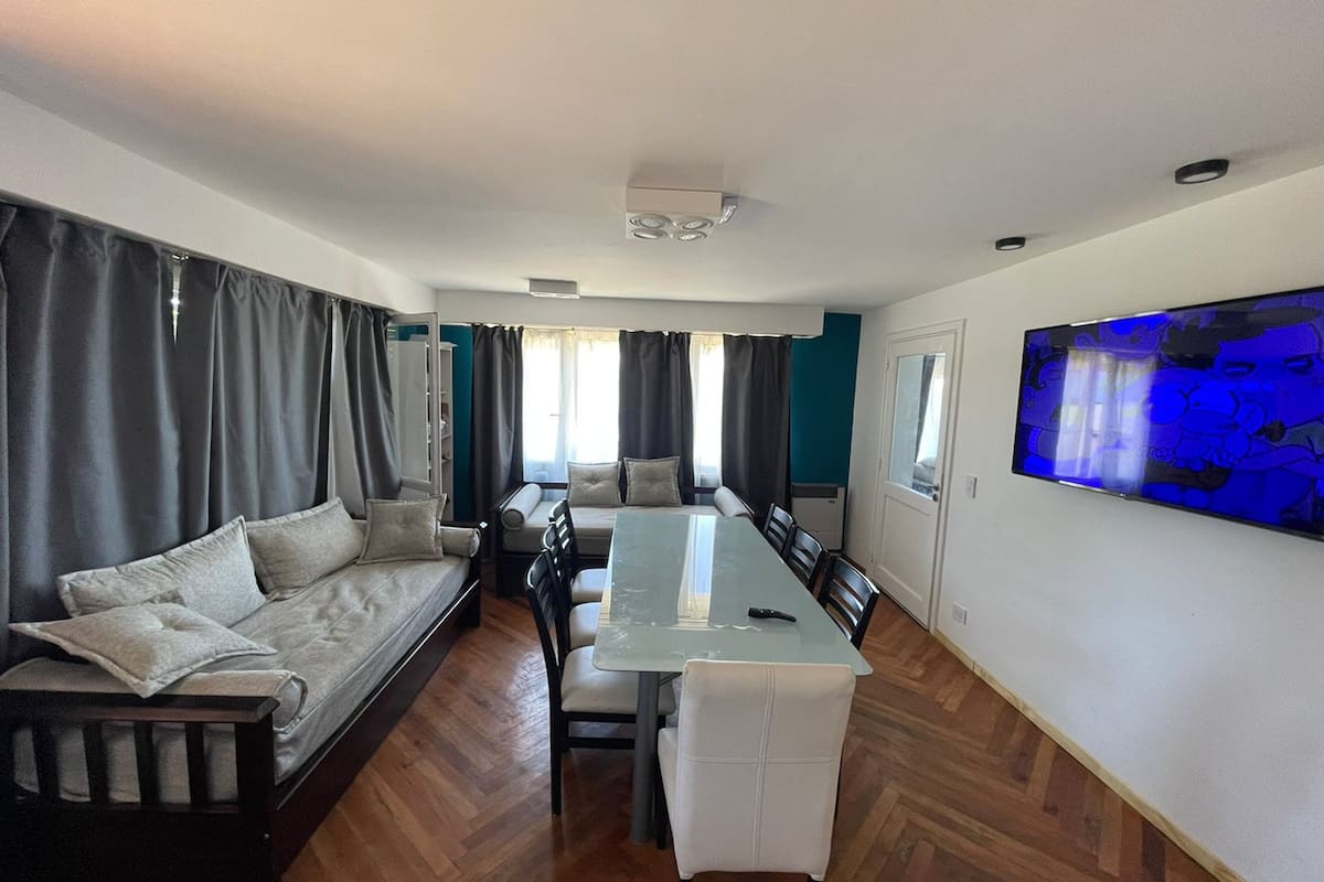 Ein modernes Wohnzimmer mit einer Couch, einem Esstisch, Stühlen, einem großen Fenster mit Vorhängen und einem am Wand montierten Fernseher.