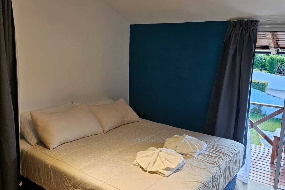 Ein Einzelbett mit beiger Bettdecke und Kissen, ein dunkles Kopfteil und Blick auf eine Terrasse sowie Grünfläche.
