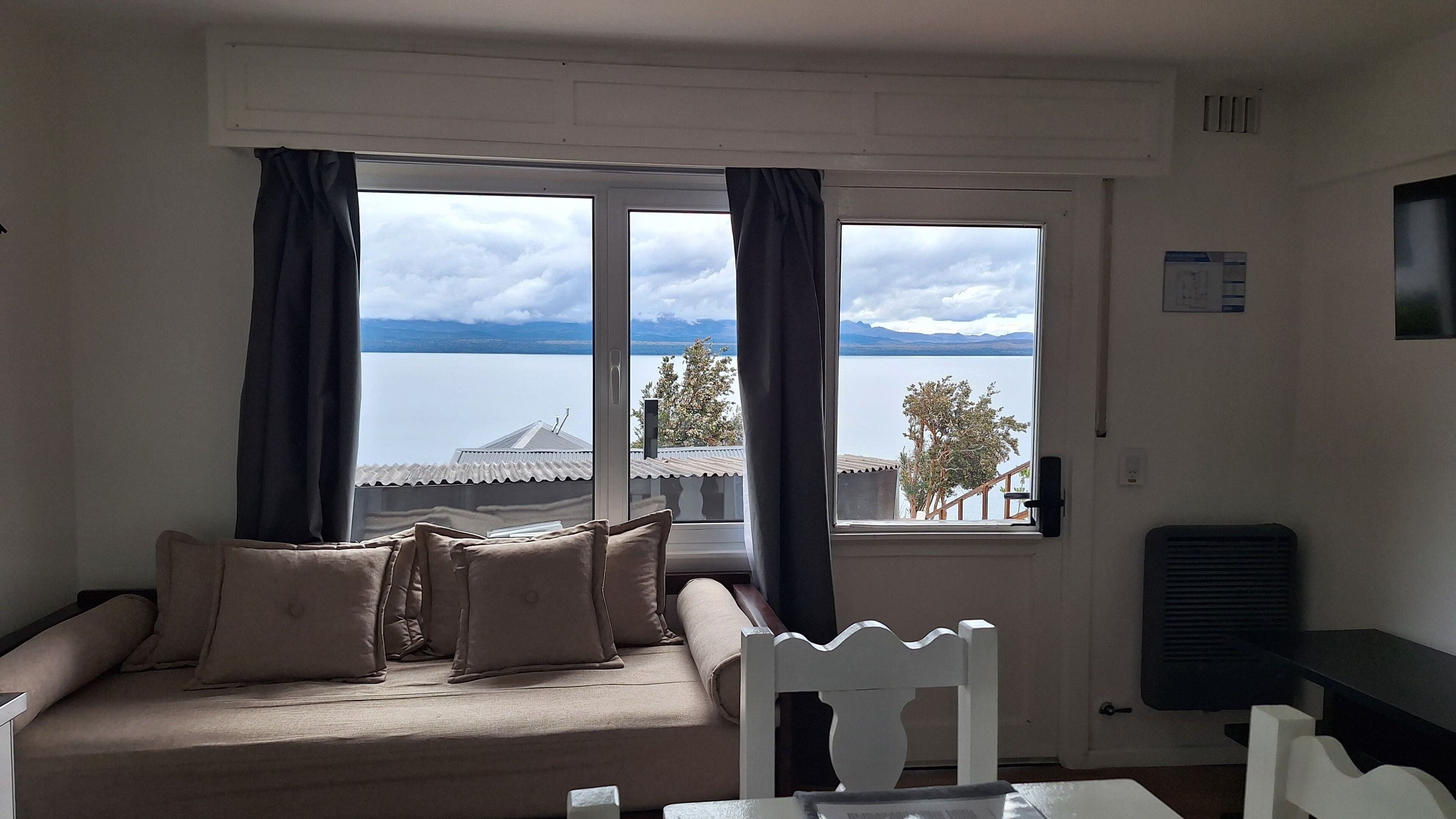 A room with a sofa and a chair, a window with curtains, and a view of a body of water and mountains.
